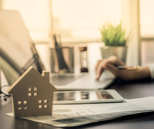 Une petite maquette de maison en bois est posée sur un bureau, devant des documents financiers et une tablette. En arrière-plan, une personne travaille sur un ordinateur portable, un crayon à la main. La scène est lumineuse et reflète un environnement de travail professionnel, suggérant la planification ou l'organisation d'un projet immobilier ou d'un investissement.