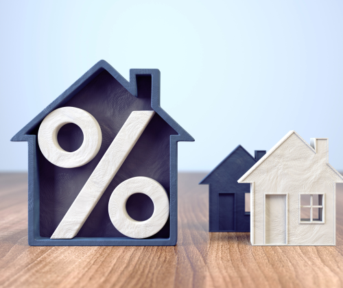 L'image montre trois maisons miniatures posées sur une surface en bois. La plus grande, de couleur bleu foncé, a un symbole de pourcentage blanc à l'intérieur, symbolisant probablement les taux d'intérêt ou des aspects financiers liés à l'immobilier. Deux autres maisons, plus petites, sont placées à côté : une blanche et l'autre bleue, représentant peut-être différentes options immobilières. L'arrière-plan est flou, mettant l'accent sur les maisons et le symbole de pourcentage.